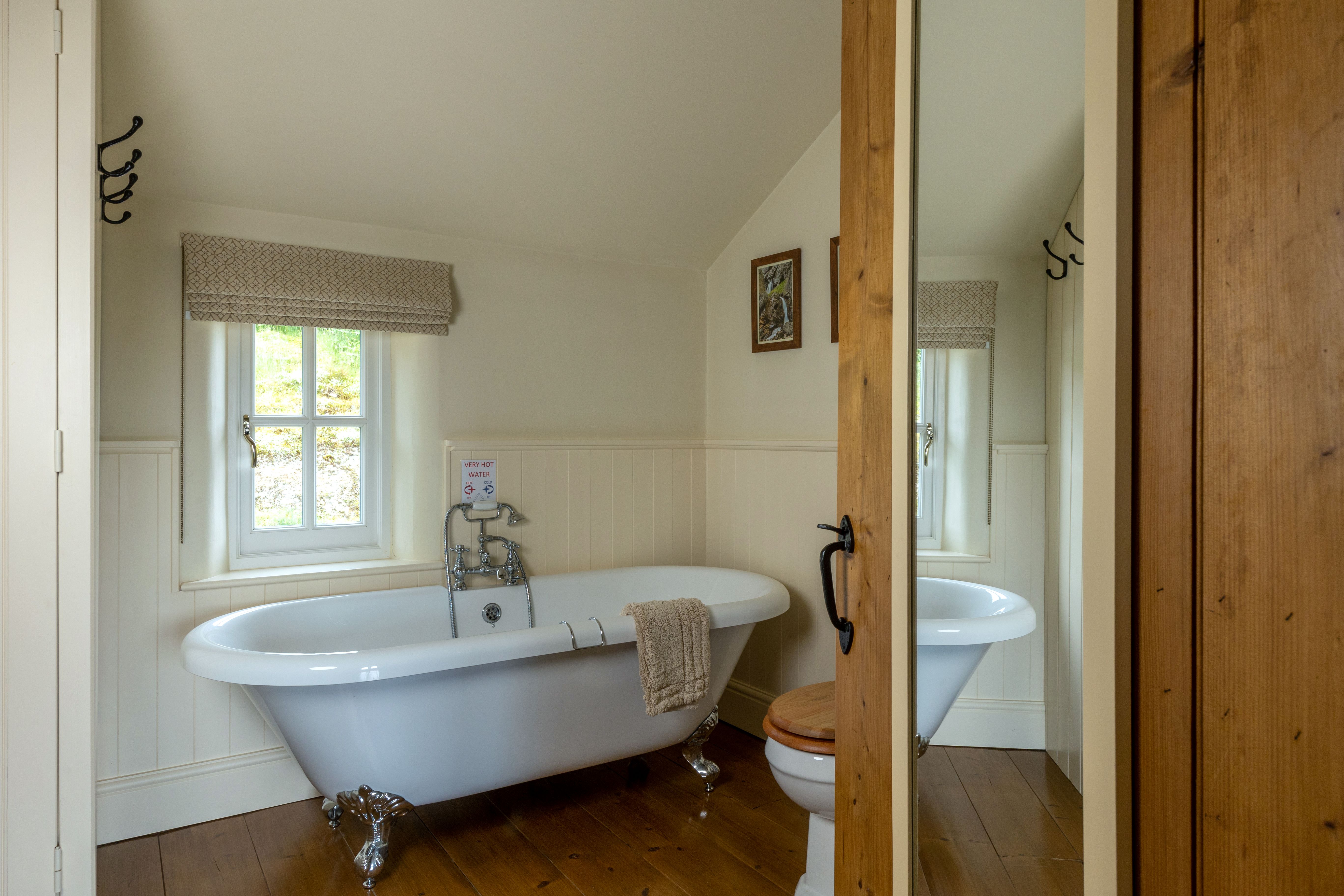 A traditional white roll top claw foot bath in a bathroom with a warm coloured wooden floor
