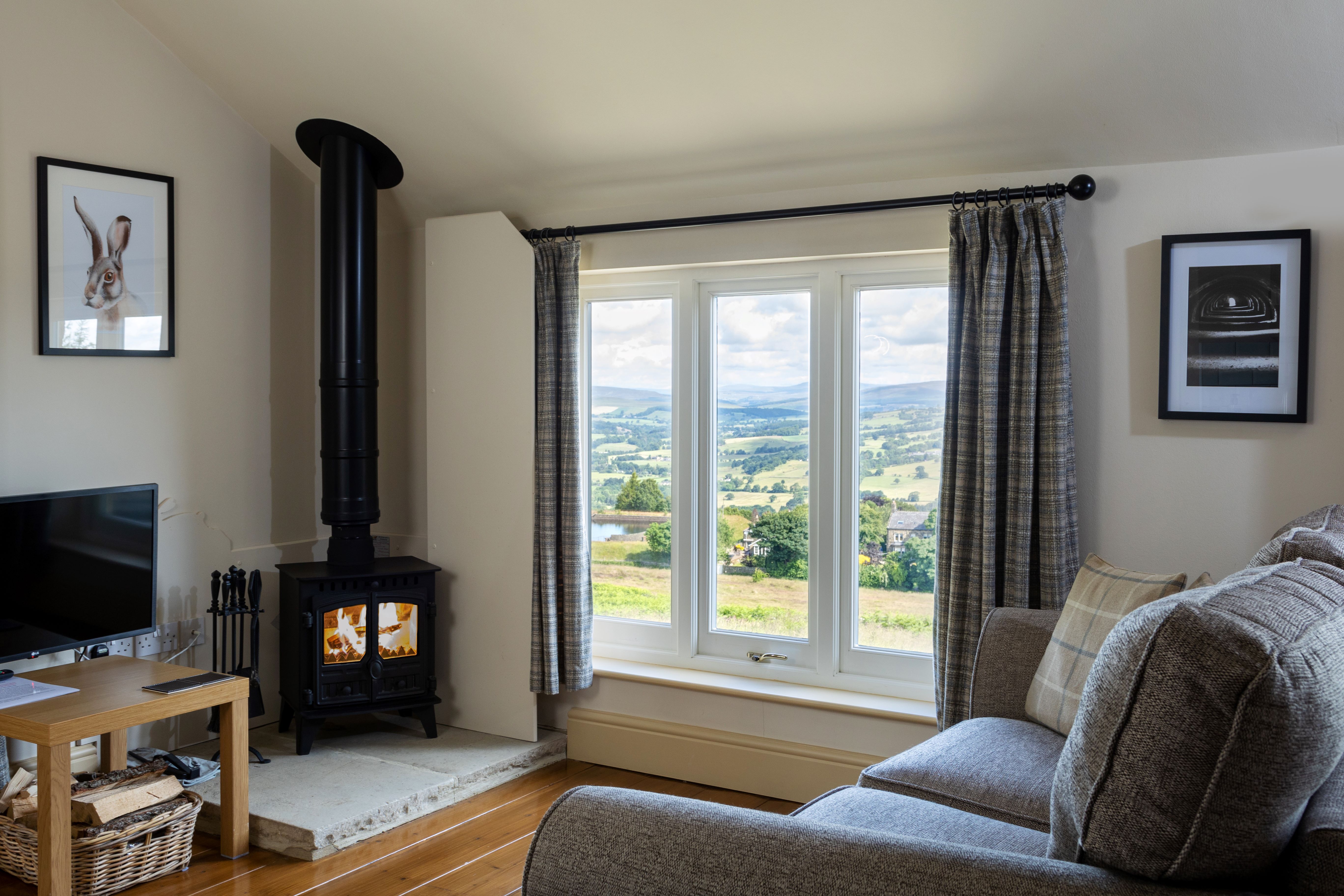 A seating area with a grey sofa, wood burner, and TV, with a view out onto rolling green hills through a large window