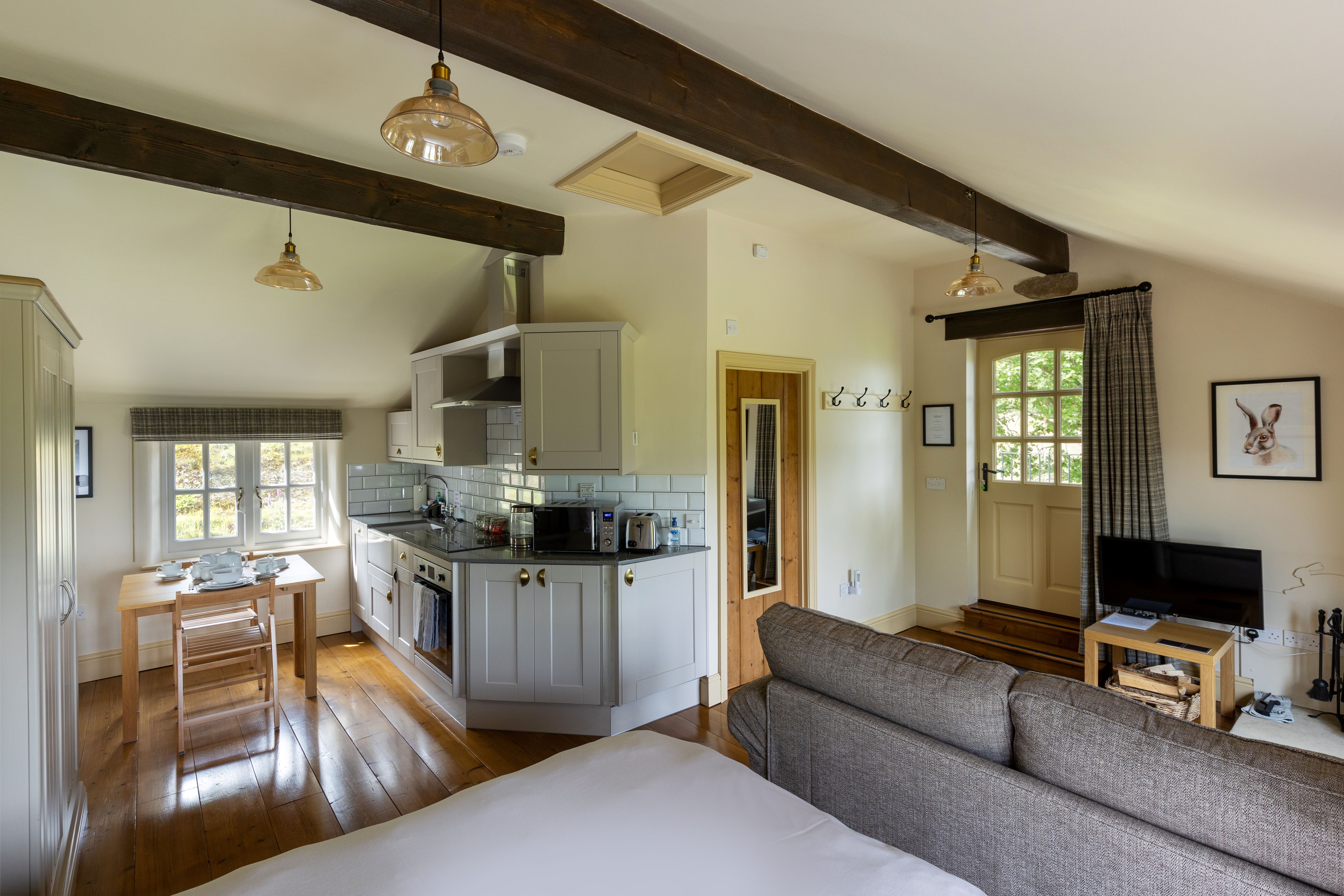 A wide angle shot of the L shaped internal layout of the studio with a kitchen and eating area to the left, the bed in the foreground, a seating area with a sofa and log burner to the right, and a door to the bathroom in the background