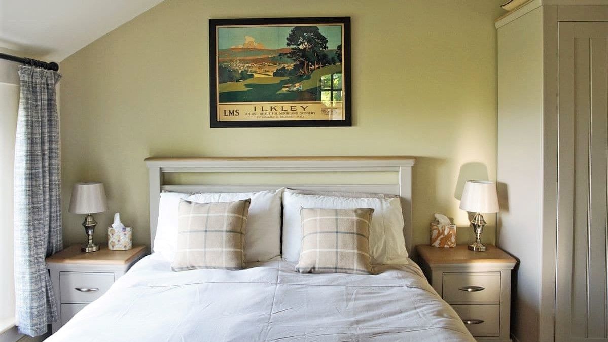 A double bed next to a window with a large retro poster for the town of Ilkley hangin on the wall above the headboard